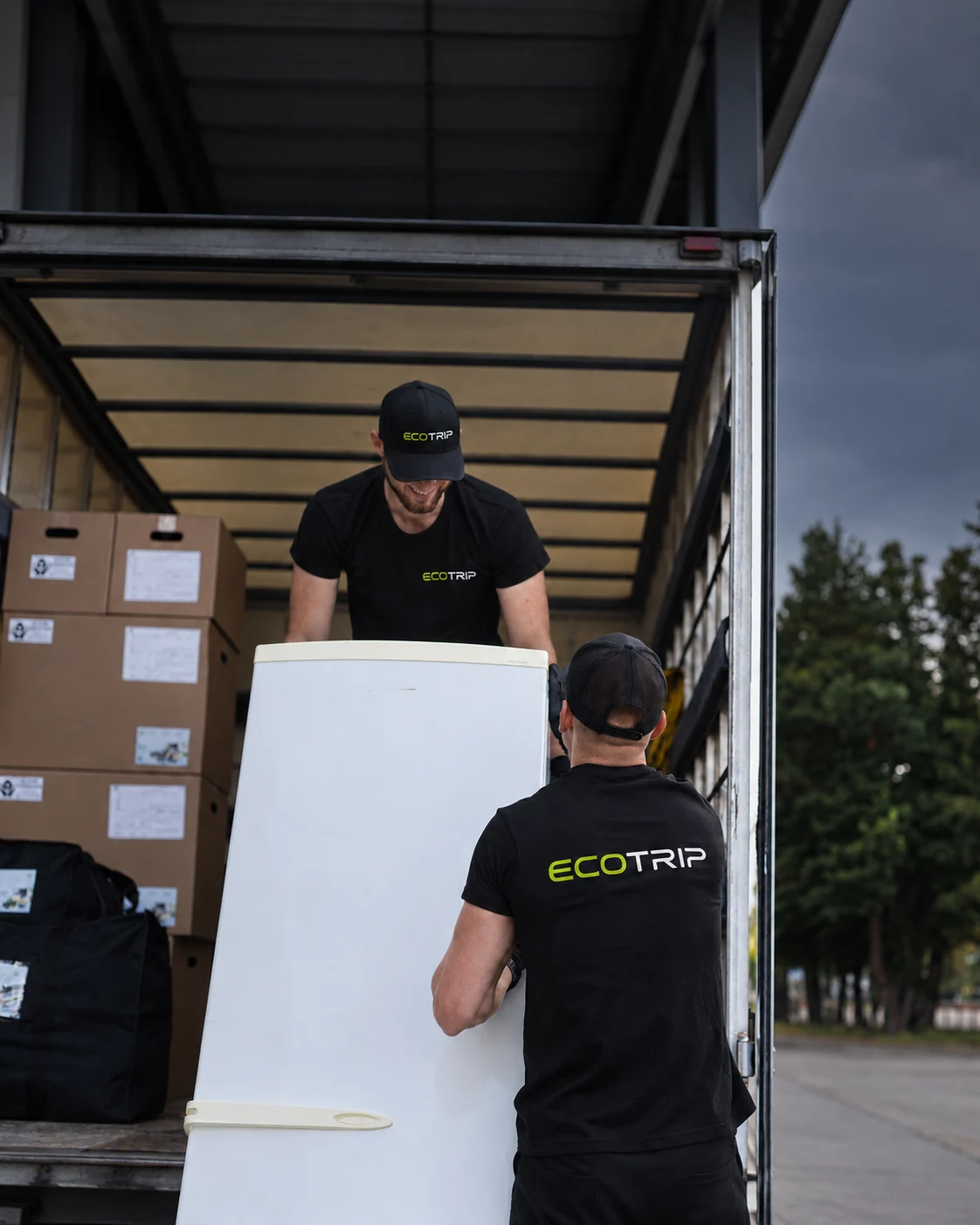 Two EcoTrip staff handing over a refrigerator at the opening of a vehicle’s cargo area. Illustration.