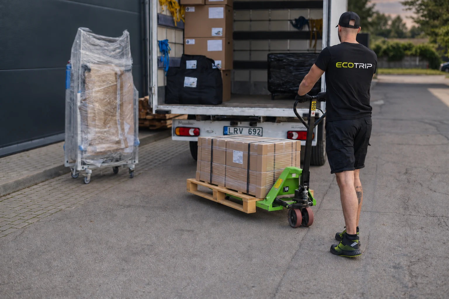 An EcoTrip staff member moving packed and secured boxes on a pallet truck beside a vehicle. Illustration.